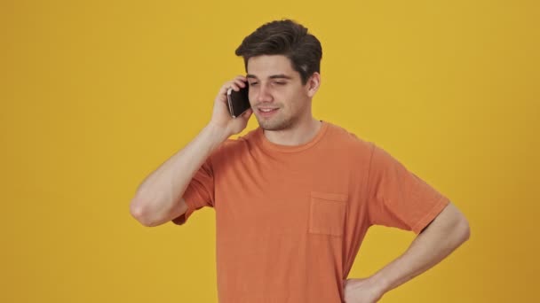 Happy handsome man wearing in t-shirt talking by smartphone and rejoicing over yellow background - Imágenes, Vídeo