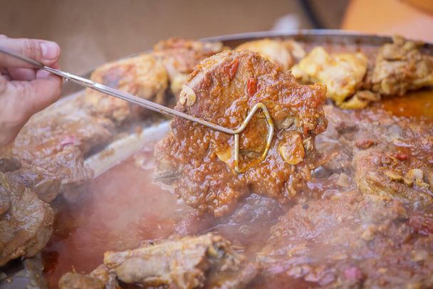 Detail of a human hand turning a pork steak meat cooked in vegetable sauce together with sausages and chicken meat on a barbeque fire in a traditional croatian kotlovina dish. - Photo, Image
