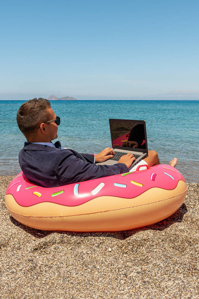 Businessman using laptop computer on inflatable donut on tropical beach. Summer vacation concept. - 写真・画像