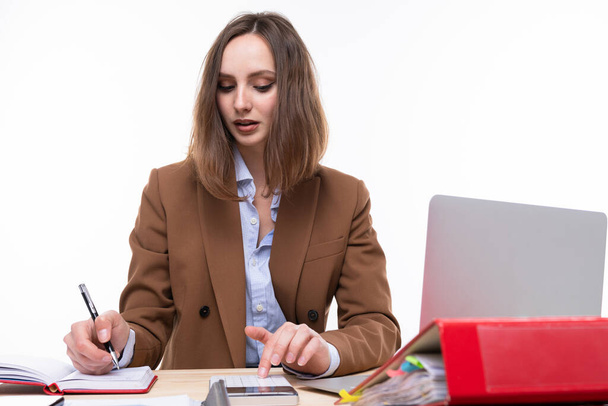 Dziewczyna w brązowym garniturze liczy na kalkulator i pisze w notatniku siedząc w miejscu pracy. Izolacja - Zdjęcie, obraz
