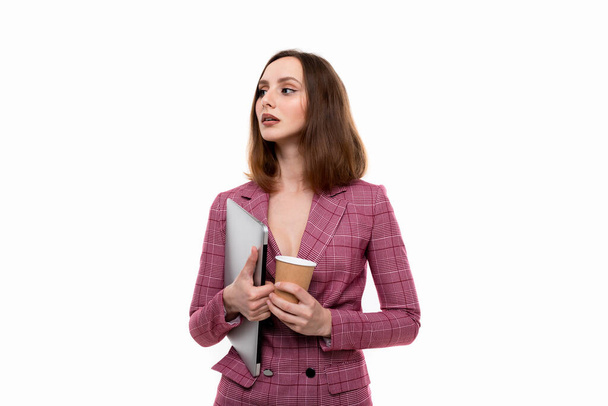 ピンクのジャケットを着た美しい女の子がノートパソコンを持ち、白い背景にコーヒーを飲みます。営業形態 - 写真・画像
