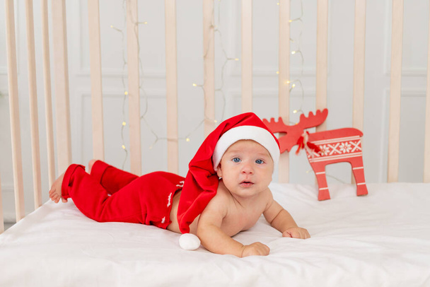 ребенок 6 месяцев в костюме Санты, лежащий в кроватке дома на животе, подарок на новый год. Рождественские праздники - Фото, изображение