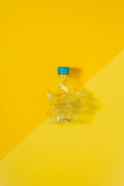 Vertical color image with an overhead view of a transparent and crushed plastic bottle with blue cap on a yellow background. Recycling and environment concept. - Photo, Image
