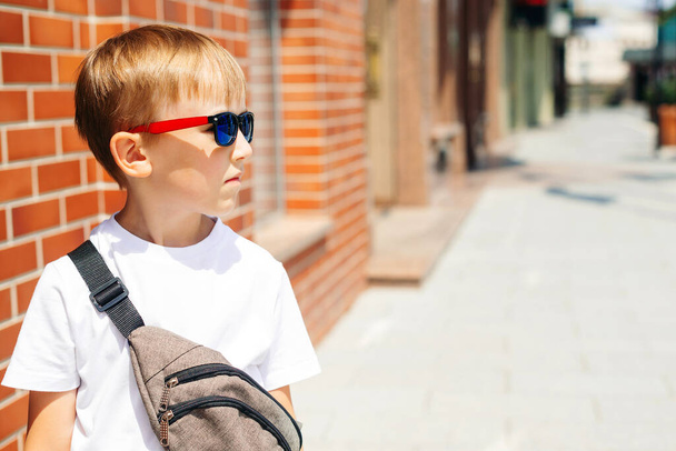 Cute kid in sunglasses at city street. Trendy boy in a white t-shirt with waist bag. Fashion handsome boy outside. Summer fashion. Happy little guy in trendy sunglasses posing outdoors. Urban style. - Фото, изображение