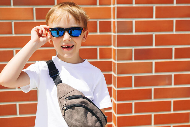 Happy little guy in trendy sunglasses outdoors. Fashion handsome boy posing over modern brick wall. Cute kid in sunglasses at city street. Trendy boy in a white t-shirt with waist bag. Summer fashion. - Фото, изображение