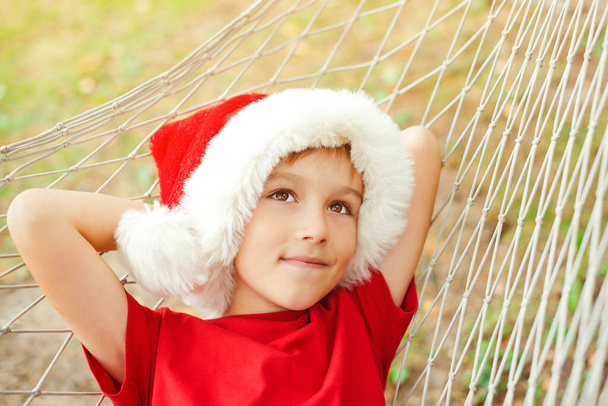 Schattig kind in kerstmuts en rustend op hangmat. Kleine jongen droomt over kerstcadeaus. Kerstvakantie met het gezin op tropisch land. Kerststemming, dromen, geschenken - Foto, afbeelding