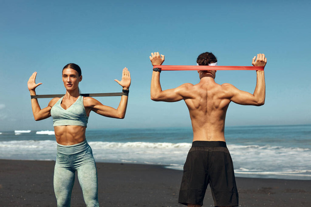 Beach. Sport Couple Workout With Resistance Band Outdoor. Handsome Man And Sexy Woman In Fashion Sporty Outfit Training On Coast. Exercising On Active Summer Vacation For Strong Muscular Body. - Foto, Imagen