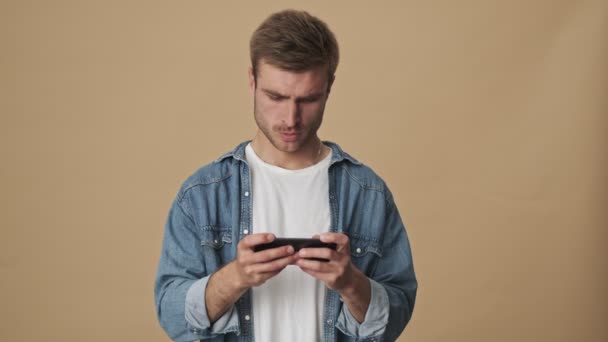 Un joven serio y concentrado está jugando un juego en su teléfono inteligente sosteniéndolo horizontalmente de pie sobre la pared beige en el estudio
 - Metraje, vídeo