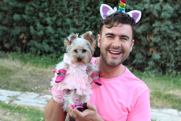 ピンクのTシャツを着たハンサムな若いゲイの男とユニコーン角で彼の子犬と庭で - 写真・画像