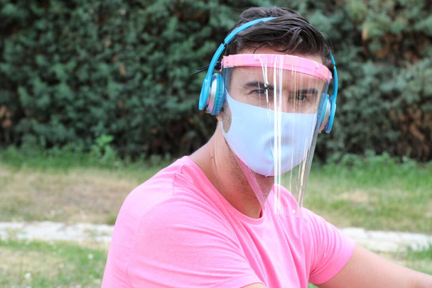 ピンクのTシャツの保護マスクと庭でヘッドフォンと顔のシールドでハンサムな若い男のクローズアップ肖像画 - 写真・画像
