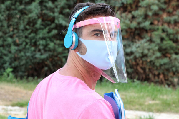 Nahaufnahme Porträt eines hübschen jungen Mannes in rosa T-Shirt Schutzmaske und Gesichtsschutz mit Kopfhörern im Garten - Foto, Bild