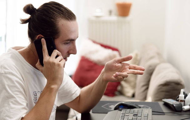 Handsome young man in casual clothing talking on the phone while sitting on the desktop in living room - 写真・画像