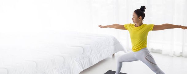 joven asiática dama vistiendo amarillo camiseta haciendo ejercicio yoga pose en el dormitorio en la mañana después de despertarse
. - Foto, Imagen