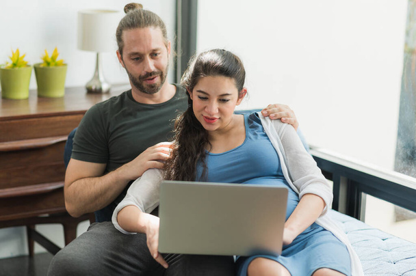 Szakállas férfi és a terhes felesége filmet néznek laptopon boldognak érzik magukat, miközben otthon ülnek a kanapén. Családi fogalom. - Fotó, kép