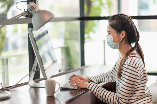 Schöne asiatische Frau mit Schutzmaske arbeiten morgens von zu Hause aus. Junge asiatische Frau arbeitet mit Computer bleiben zu Hause. - Foto, Bild