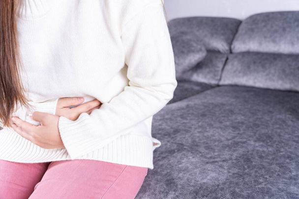 Jeune femme souffrant de fortes douleurs abdominales ou menstruations tout en étant assis sur le canapé à la maison. Soins de santé concept médical ou de la vie quotidienne. - Photo, image
