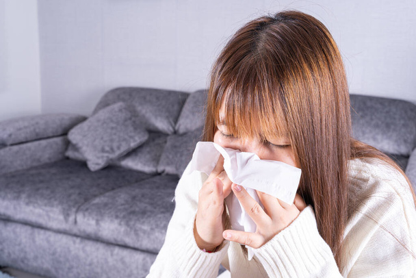 Asiatische schöne Frau bläst eine Nase zu Gewebe, während sie zu Hause auf dem Sofa sitzt. Gesundheitswesen medizinisches oder tägliches Lebenskonzept. - Foto, Bild