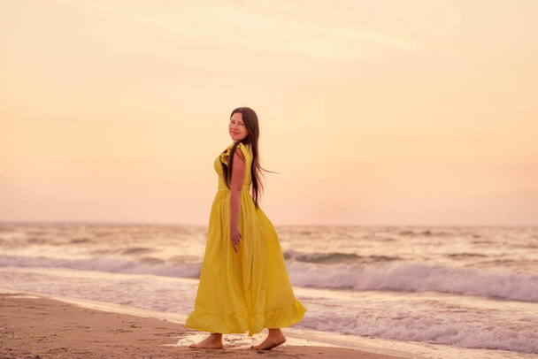 A girl in a long dress with long black hair walks at dawn along a deserted beach along the line of the surf.  - Foto, immagini