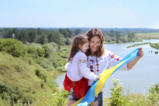 Feliz mamá ucraniana sonriente y su hija en vyshyvanka (camisas bordadas) con una bandera amarillo-azul sobre un fondo de montañas y un río.Día de la Independencia de Ucrania. Día Nacional de la Bandera de Ucrania
 - Foto, imagen