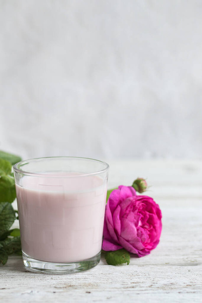 Ayurvedic drink pink milk or matcha. A glass of refreshing cocktail stands on a wooden white background. Nearby lies a lilac rose. Copy space - Foto, imagen