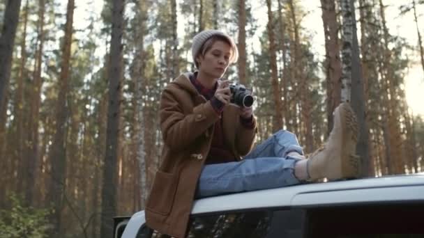 Jonge blanke vrouw in muts zittend op autodak op bosweg en met camera foto 's van de natuur in de herfst - Video