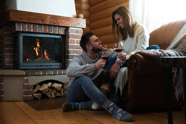 Lovely couple with glasses of wine near fireplace indoors. Winter vacation - Foto, Imagem