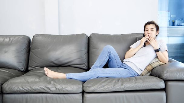 Joven mujer asiática relajándose en casa viendo la televisión y cambiando el canal con un control remoto
 - Foto, Imagen