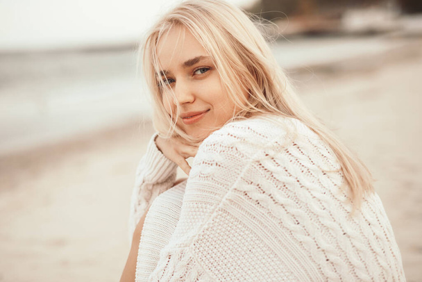 Smiling blonde woman in warm sweater sitting on beach - Фото, зображення