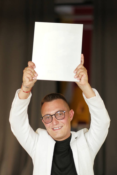 A young man of 30-35 years old in a white jacket, black turtleneck, glasses and a short haircut holds an empty sign in his hands - Foto, Bild