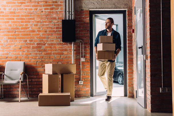 hombre guapo sosteniendo cajas de cartón mientras camina en la nueva oficina
  - Foto, Imagen