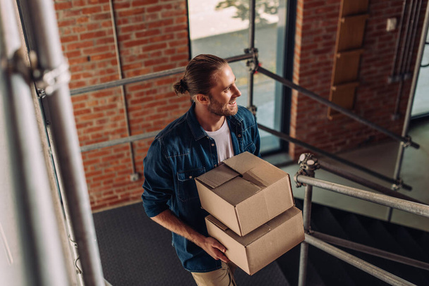 selective focus of smiling businessman holding boxes and walking on stairs in new office - Fotoğraf, Görsel