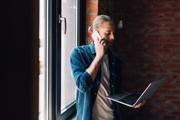 selective focus of businessman looking at laptop and talking on smartphone near window  - Φωτογραφία, εικόνα