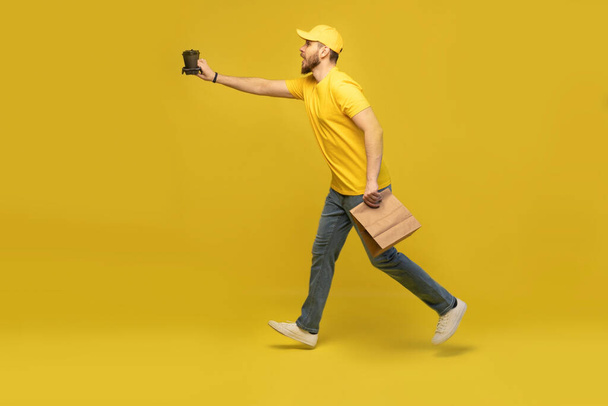 Portrait of young delivery man in yellow uniform with paper packet and takeaway coffee isolated over yellow background. - Φωτογραφία, εικόνα