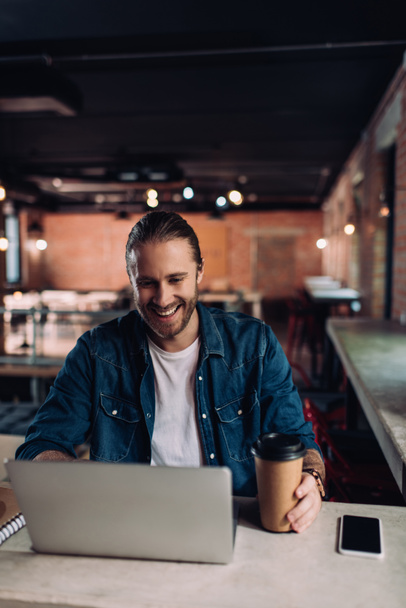boldog üzletember néz laptop és megható papír pohár közelében okostelefon üres képernyő - Fotó, kép