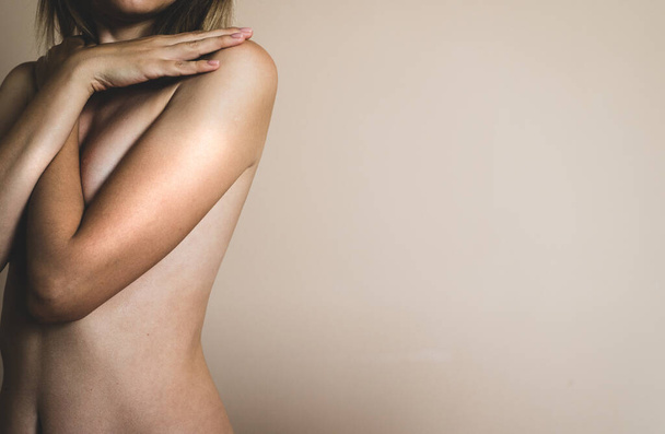 Una giovane donna bionda nuda incrocia le braccia sul petto su uno sfondo beige - Foto, immagini