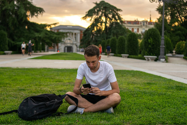 Foto de un hombre joven y atractivo con una camiseta blanca sentada en la hierba revisando su teléfono - Foto, Imagen