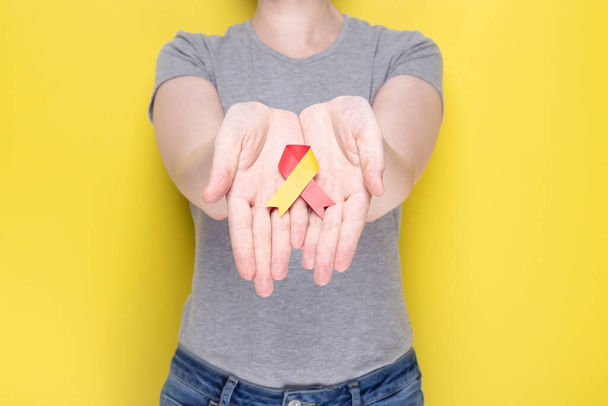 World Hepatitis Day concept. Girl in gray T-shirt holds in her hands awareness symbol red-yellow ribbon. Yellow background - Фото, зображення