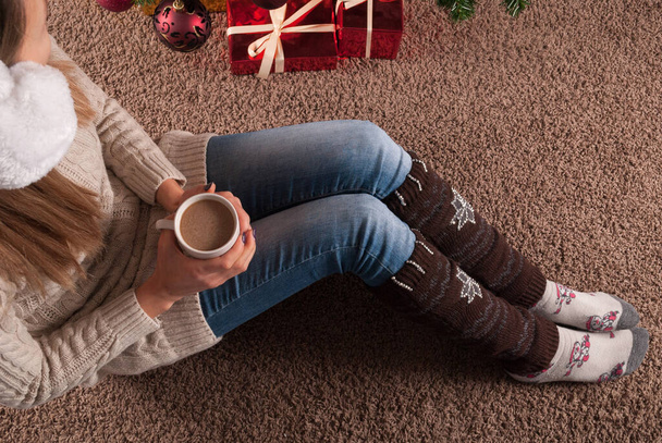 Молода дівчина сидить на підлозі на килимі з чашкою кави в руці і поруч з різдвяною подарунковою коробкою, крупним планом
 - Фото, зображення