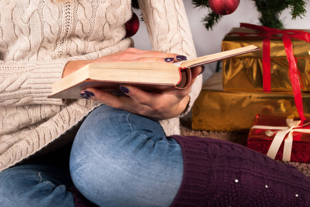 Girl in sweater holding book in hands and Christmas tree and gift box in background, close up, selective focus - Photo, Image