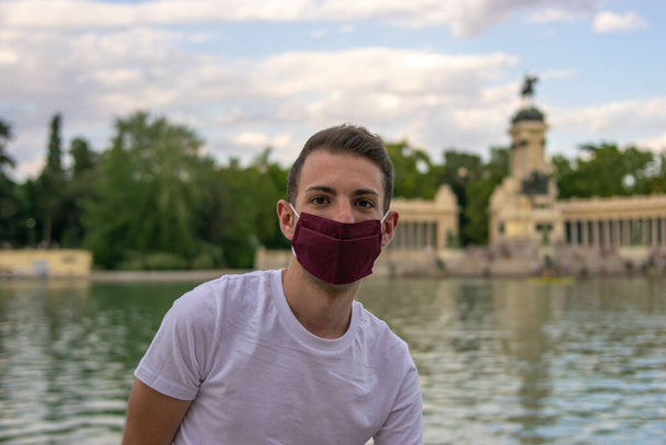 Foto de um homem jovem e atraente vestindo uma máscara facial e sentado ao lado de um parque bonito e graan isolado na natureza - Foto, Imagem