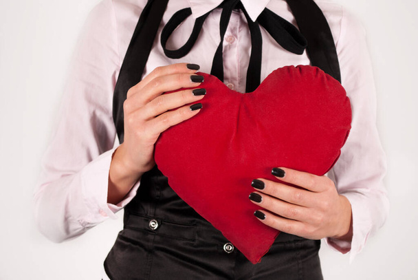 Rotes Herzkissen in Frauenhand isoliert auf weißem Hintergrund. Frau ist elegant und mit Krawatte und schwarzen Nägeln. Nahaufnahme - Foto, Bild