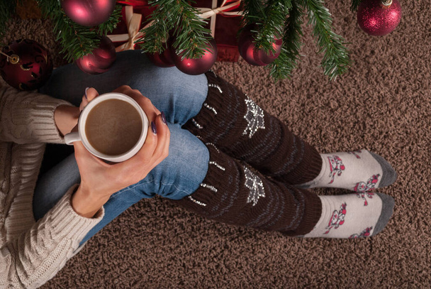 Femme assise sur le sol et réchauffe la prise de jambe et avec une tasse de café à la main et à côté de l'arbre de Noël, gros plan, vue sur le dessus - Photo, image