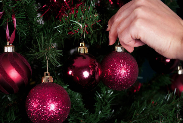 Lány kéz lóg piros dekoráció karácsonyfa és elmosódott fa a háttérben. Közelről. - Fotó, kép
