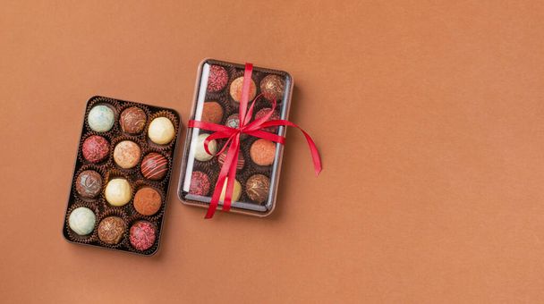 Chocolate sweets from various chocolate toppings in a box with a satin ribbon bow dark brown background. The concept of a festive dessert. Horizontal frame copy space. Top view flat layout - 写真・画像