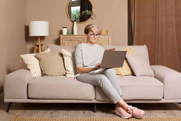 Young female freelancer working or learning from home.  Pretty intelligent woman in casual clothes sitting on a sofa typing on a laptop front view. - Фото, изображение