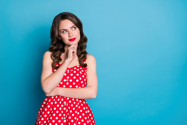 Photo of attractive curly lady minded good mood arms crossed look up empty space interested creative person arm on chin wear red dotted retro dress isolated blue color background - Foto, afbeelding