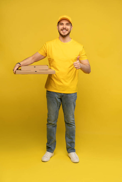 Portrait of young delivery man in yellow uniform with pizza boxes isolated over yellow background. - Fotoğraf, Görsel