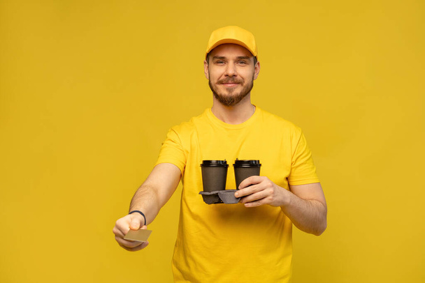 Portrait of young delivery man in yellow uniform with takeaway coffee isolated over yellow background. - Foto, Imagem