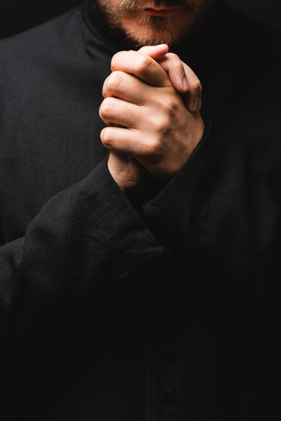 καλλιεργημένη άποψη του ιερέα με σφιγμένα χέρια προσεύχεται απομονωμένη σε μαύρο  - Φωτογραφία, εικόνα