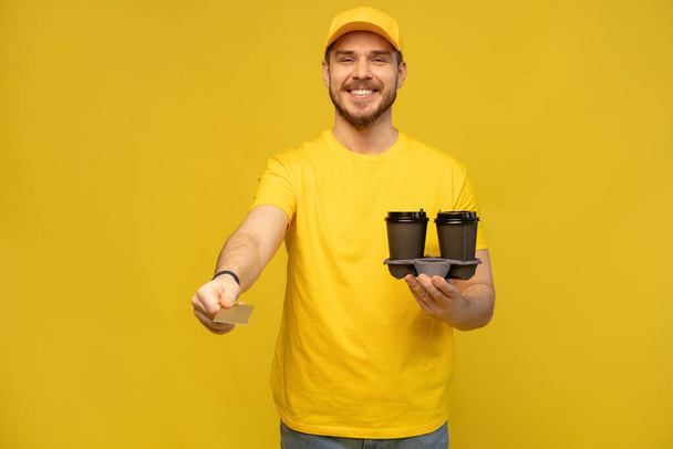 Portrait of young delivery man in yellow uniform with takeaway coffee isolated over yellow background. - Photo, image